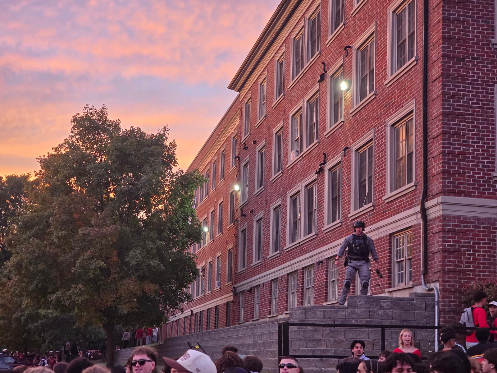 A police officer on the steps of a brick residence at sunset, a crowd of students below