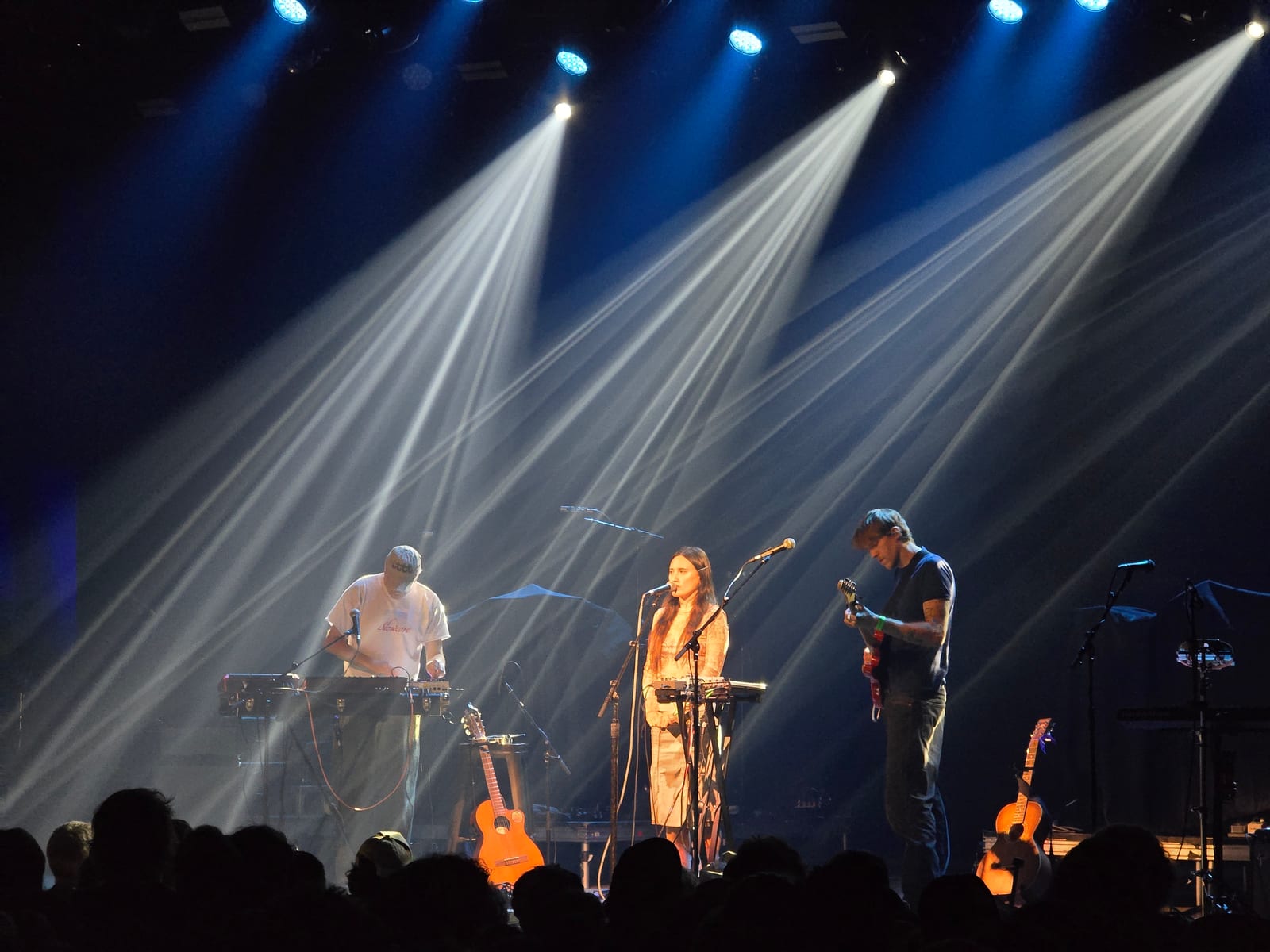 A three piece band on a hazy stage under crossing white beams