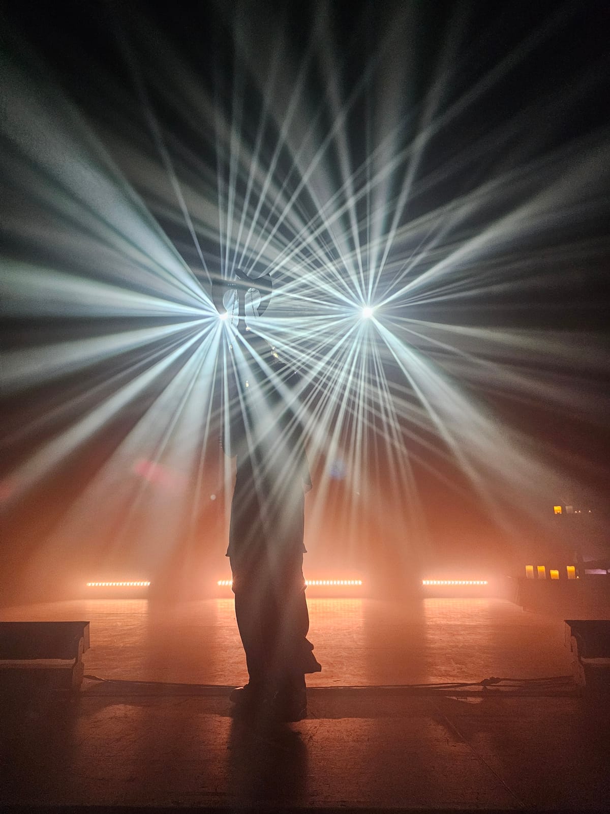 A performer silhouetted on a hazy stage between two starburst spotlights