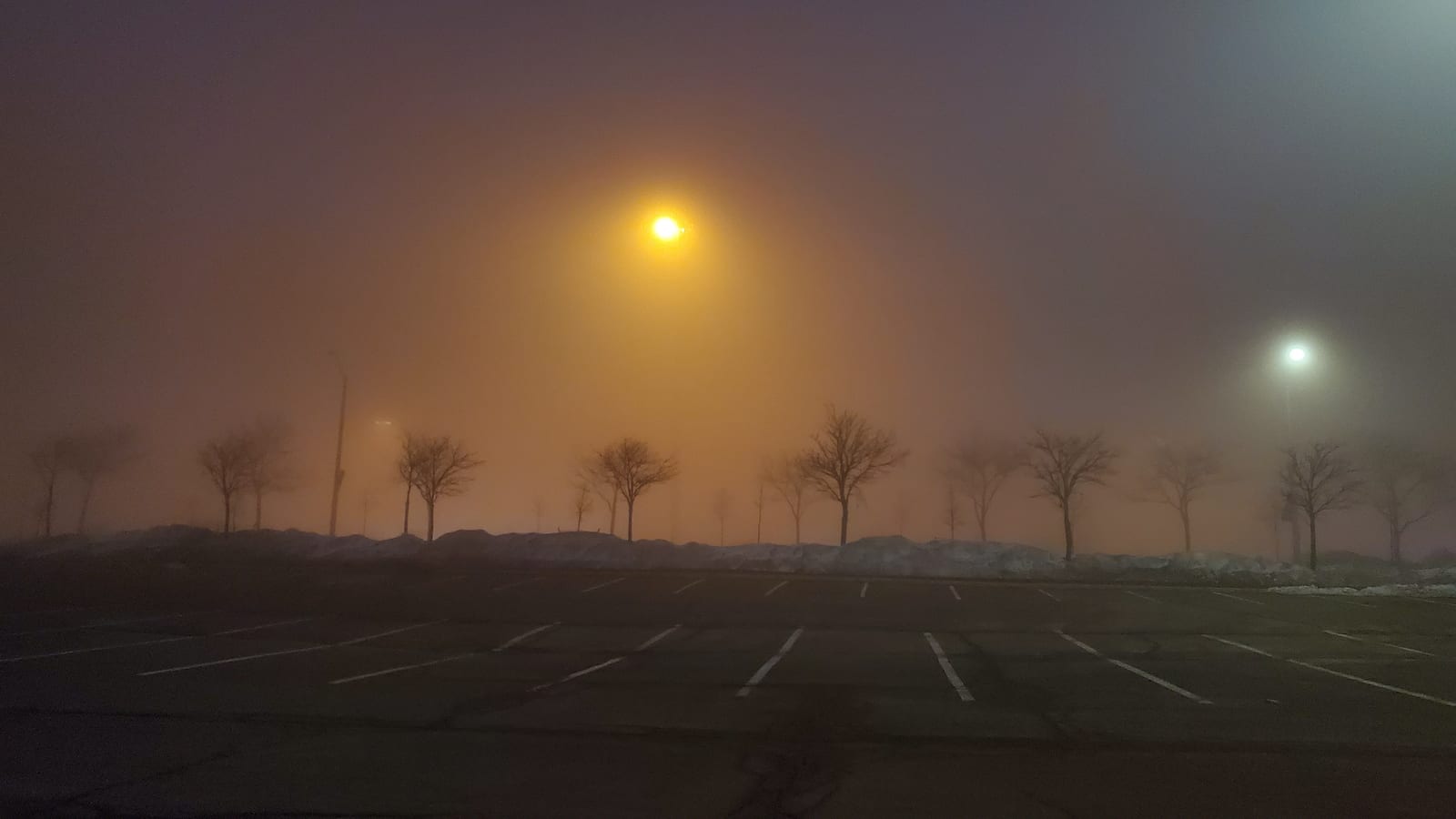An empty parking lot in thick night fog, a sodium streetlight glowing orange over bare trees and snowbanks